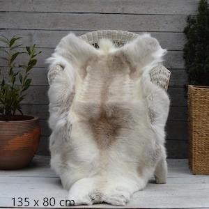 May include: A light-coloured reindeer hide rug, measuring 135 x 80 cm, is draped over a white wicker chair. The rug features a mix of white, cream, and brown tones, with a soft, textured appearance. The rug is displayed on a wooden surface.