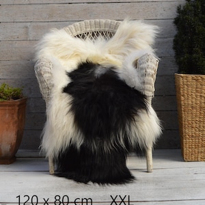 May include: A long-haired sheepskin rug featuring a two-tone design with a large black centre section and cream-coloured edges. The rug is draped over a white wicker chair and includes the text 120 x 80 cm XXL printed at the bottom of the image.