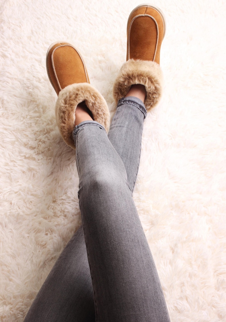 Op de afbeelding: Een paar bruine su&egrave;de pantoffels met een rand van nep bont, gedragen door iemand die grijze jeans draagt. De pantoffels staan op een wit, pluche tapijt.