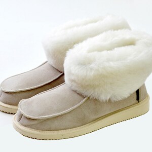 Op de afbeelding: Een paar beige su&egrave;de pantoffels met witte nep bont trim. De pantoffels hebben een afgeronde neus en een witte rubberen zool.