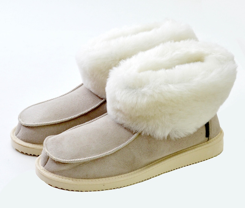 Op de afbeelding: Een paar beige su&egrave;de pantoffels met witte nep bont trim. De pantoffels hebben een afgeronde neus en een witte rubberen zool.