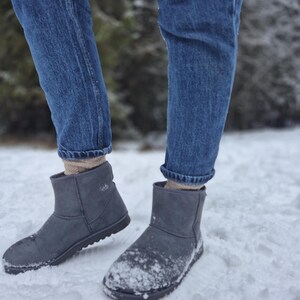 May include: A pair of gray suede ankle boots with a black sole. The boots are worn by a person wearing blue jeans and standing in the snow.