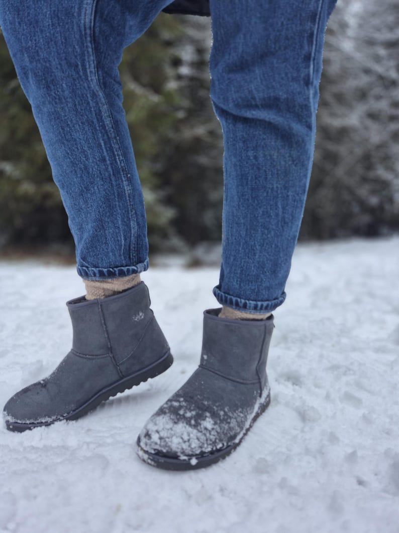 May include: A pair of gray suede ankle boots with a black sole. The boots are worn by a person wearing blue jeans and standing in the snow.
