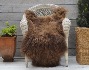 Op de afbeelding: Een bruin schapenvachtkleed, XL-formaat, gedrapeerd over een witte rotan stoel. Het kleed meet 105 x 70 cm. De harige textuur van het kleed contrasteert met de gladde stoel. De achtergrond is een houten muur en een plant in een pot.