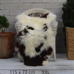Pode incluir: Uma pele de ovelha de pelo comprido com um padrão manchado de tons branco-creme e castanho-escuro. A pele está colocada sobre uma cadeira de vime branca e inclui o texto impresso 115 x 73 cm XXL na parte inferior da imagem.