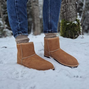 May include: A pair of brown suede boots with a sheepskin lining. The boots are worn by a person standing in the snow.