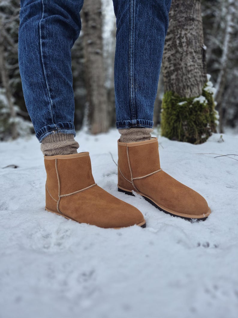 May include: A pair of brown suede boots with a sheepskin lining. The boots are worn by a person standing in the snow.