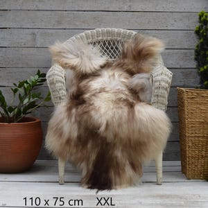 Könnte beinhalten: Ein langhaariges, flauschiges Schaffell mit einer Mischung aus Creme-, Beige- und Dunkelbrauntönen. Das Fell liegt über einem weißen Korbstuhl und ist mit den Maßen 110 x 75 cm sowie der Größenbezeichnung XXL versehen.