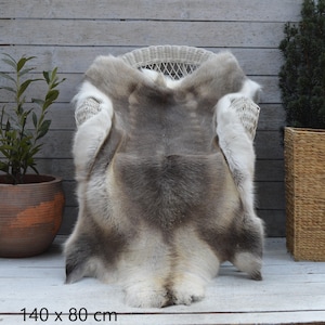Puede incluir: Una alfombra de piel de reno, de 140 x 80 cm, está colocada sobre una silla de mimbre blanca. La alfombra presenta una mezcla de pelo gris, marrón y blanco. Las dimensiones están impresas en la parte inferior de la imagen.