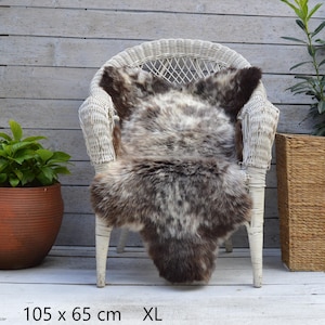 Könnte beinhalten: Ein gesprenkeltes braun-grau-weißes Schaffell mit langem, dichtem Flor. Das Fell liegt über einem weißen Korbstuhl. Der Text am unteren Bildrand gibt die Maße mit 105 x 65 cm und die Größe mit XL an.