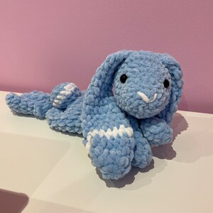 May include: A light blue crocheted bunny plush toy with white accents. The bunny is lying on its side with its head turned towards the camera.