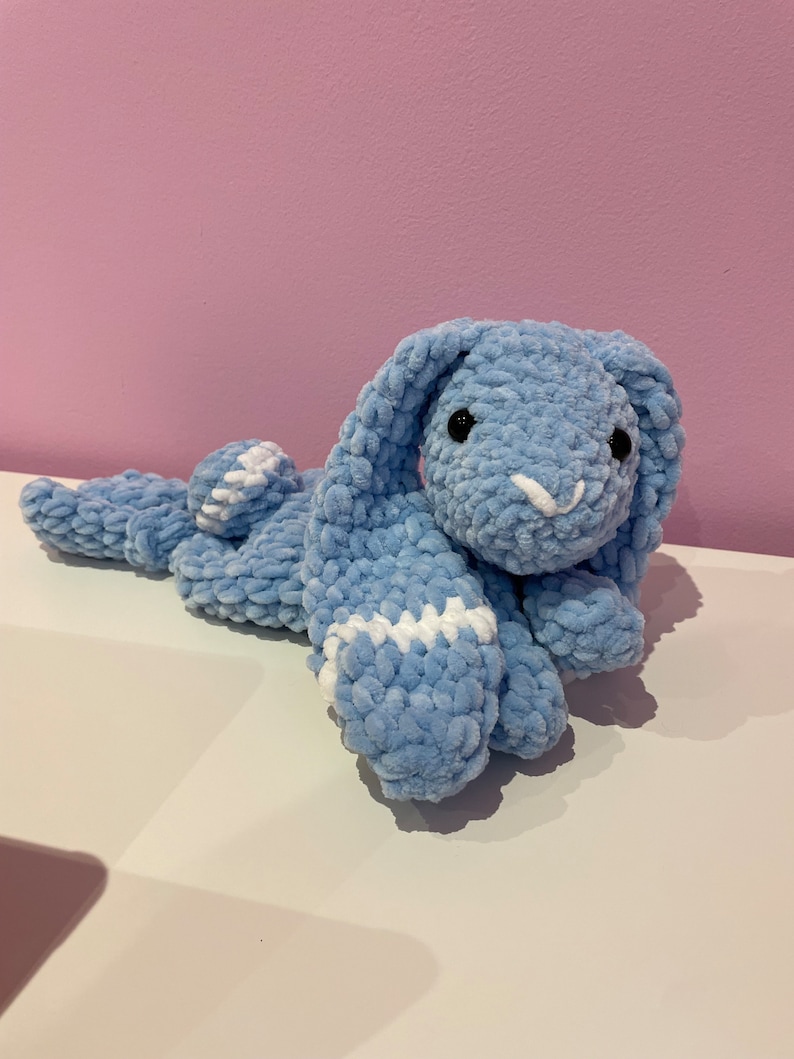 May include: A light blue crocheted bunny plush toy with white accents. The bunny is lying on its side with its head turned towards the camera.