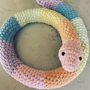 May include: A colourful crocheted snake toy in shades of pink, purple, yellow, and blue. The snake is curled into a circle shape.