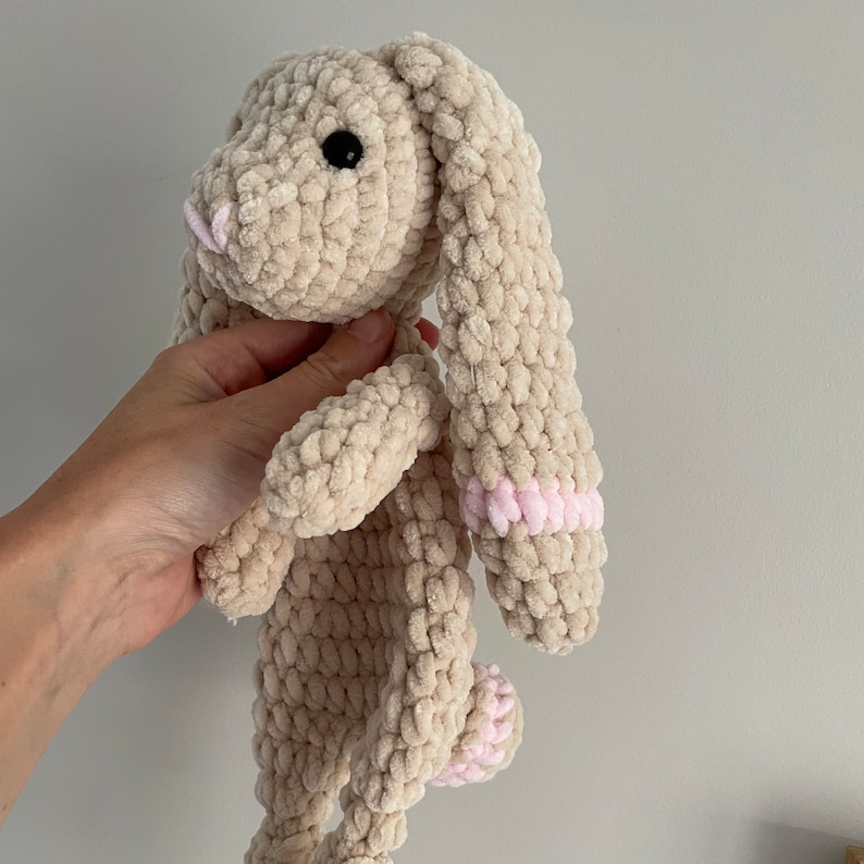 May include: A crocheted plush bunny toy with long ears. The bunny is beige with pink accents on the ears.