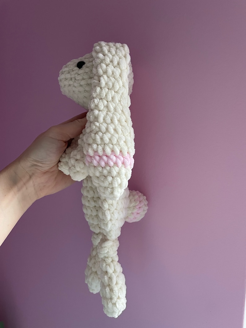 May include: A white crocheted stuffed animal shaped like a dog with pink accents. The dog is standing on its hind legs with its front paws raised.