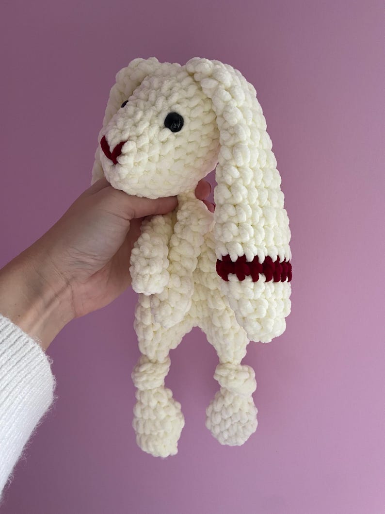 May include: White crocheted bunny with long floppy ears and a red stripe on one ear. The bunny has a small black nose and black eyes.