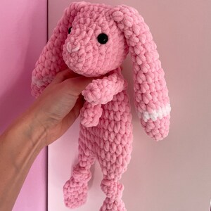 May include: A pink crocheted bunny toy with white accents on the ears. The bunny has a long body and long, floppy ears.