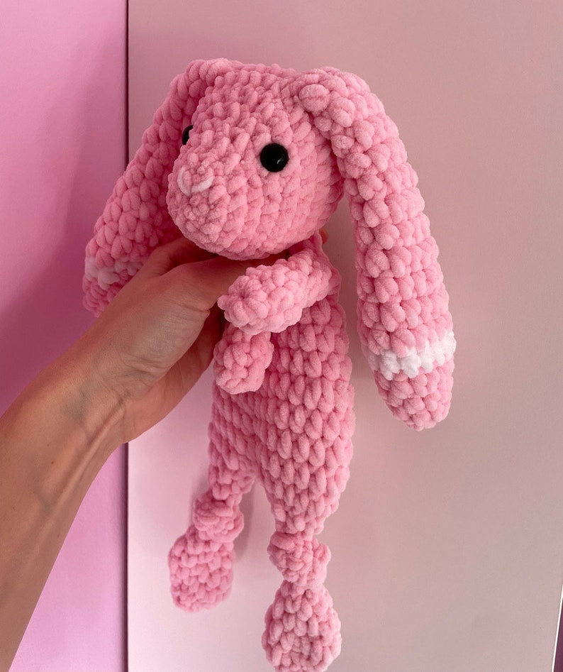 May include: A pink crocheted bunny toy with white accents on the ears. The bunny has a long body and long, floppy ears.