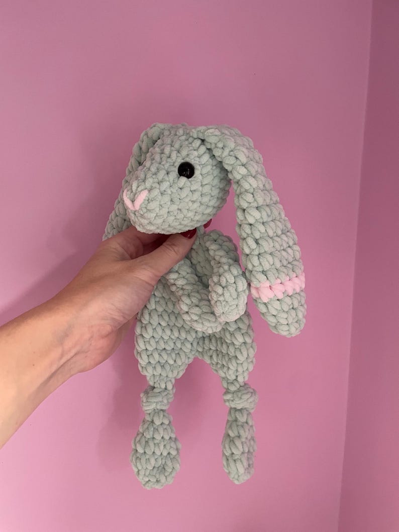 May include: A soft, green crocheted bunny toy with long ears and pink accents. The bunny has black eyes and a white nose.