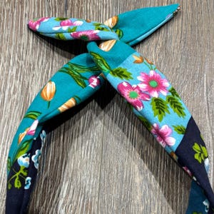 May include: A teal fabric headband with a knotted top. The headband features floral patterns in pink, white, and blue, with accents of orange and green. The headband is approximately 2.5 cm wide.