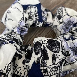 Peut inclure: Un chouchou en tissu blanc avec un imprimé de crâne noir et blanc et de fleurs. Le chouchou a un accent bleu marine.