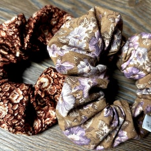Peut inclure: Deux chouchous à motifs floraux marrons et beiges. Les chouchous sont en tissu et ont un aspect plissé. Le chouchou de gauche a un motif floral brun foncé, tandis que le chouchou de droite a un motif floral brun clair avec des fleurs violettes. Le chouchou de droite porte une étiquette blanche avec le texte "The Brown Tulip" dessus.