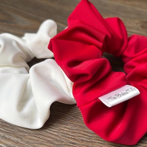 May include: Two scrunchies, one white and one red, sit on a wooden surface. The red scrunchie has a small white tag that reads "The Bridal Tale". The scrunchies are made of a soft, silky material.