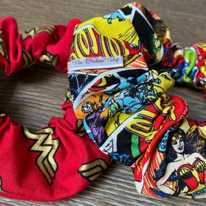 May include: Two red and yellow fabric scrunchies. One scrunchie has a Wonder Woman logo pattern. The other scrunchie has a comic book pattern with the text "The Broken Tulip".
