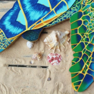 May include: A blue and green painted wooden turtle shell with yellow accents. The shell is on a sandy surface with seashells.