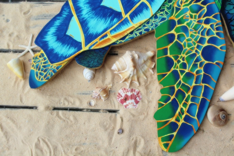 May include: A blue and green painted wooden turtle shell with yellow accents. The shell is on a sandy surface with seashells.