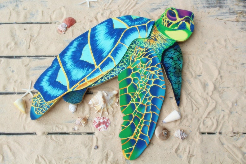 May include: A blue and green painted wooden sea turtle sculpture with a detailed shell and flippers. The turtle is lying on a sandy surface with seashells.