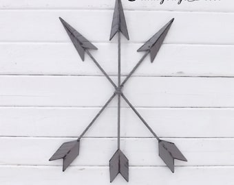 May include: A wall decor featuring five gray metal arrows arranged in a starburst pattern. The arrows are crossed in the center and point outwards. The background is a white wood panel.