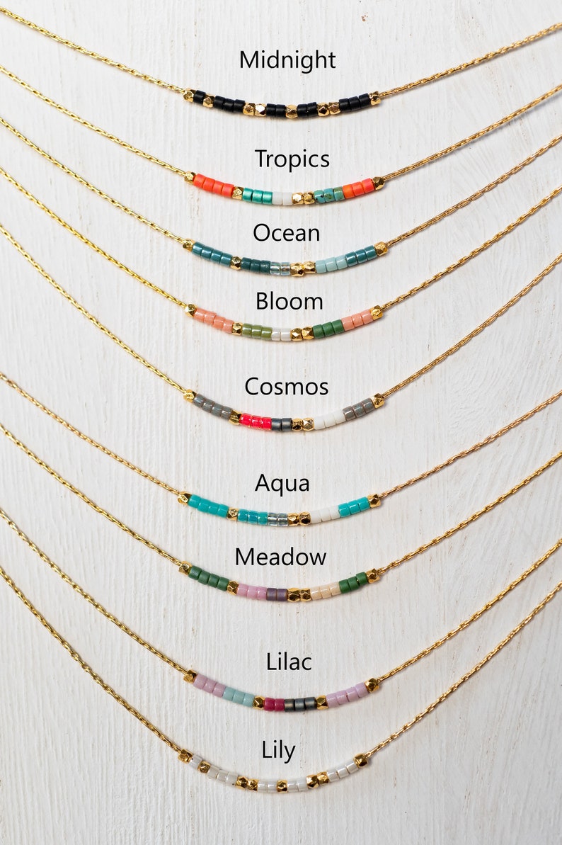 Puede incluir: Nueve collares de oro con cuentas de diferentes colores, cada collar est&aacute; etiquetado con un nombre: Medianoche, Tr&oacute;picos, Oc&eacute;ano, Floraci&oacute;n, Cosmos, Aqua, Prado, Lila, Lirio.