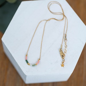 Puede incluir: Un collar de cadena dorada con una fila delicada de cuentas de colores en tonos verdes, rosas y blancos. El collar tiene un dije en forma de estrella y un cierre de langosta.