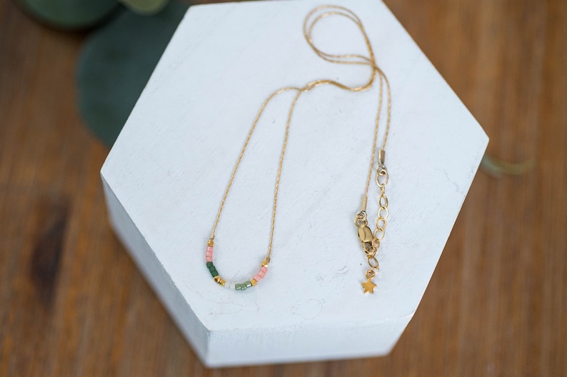 Puede incluir: Un collar de cadena dorada con una fila delicada de cuentas de colores en tonos verdes, rosas y blancos. El collar tiene un dije en forma de estrella y un cierre de langosta.