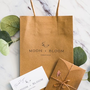Puede incluir: Una bolsa de papel marr&oacute;n con el logotipo "MOON + BLOOM CREATIONS" impreso en la parte delantera. Una peque&ntilde;a tarjeta blanca con el mismo logotipo y el texto "Gracias" es visible. Un sobre marr&oacute;n con una flor rosa y una cuerda dorada tambi&eacute;n es visible.