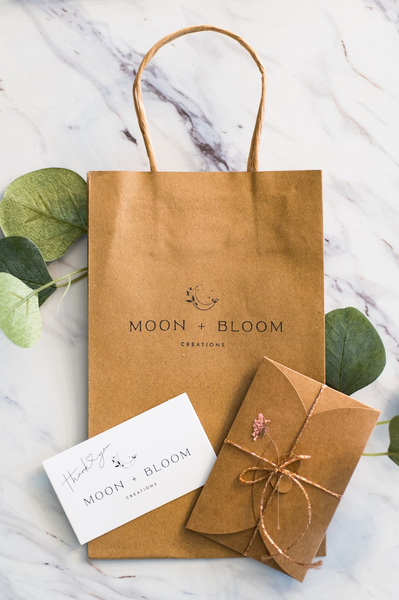 Puede incluir: Una bolsa de papel marr&oacute;n con el logotipo "MOON + BLOOM CREATIONS" impreso en la parte delantera. Una peque&ntilde;a tarjeta blanca con el mismo logotipo y el texto "Gracias" es visible. Un sobre marr&oacute;n con una flor rosa y una cuerda dorada tambi&eacute;n es visible.