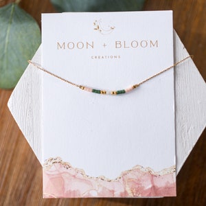 Puede incluir: Un delicado collar de cadena dorada con peque&ntilde;as cuentas rosas y verdes, presentado en una tarjeta blanca con el texto "MOON + BLOOM CREATIONS".