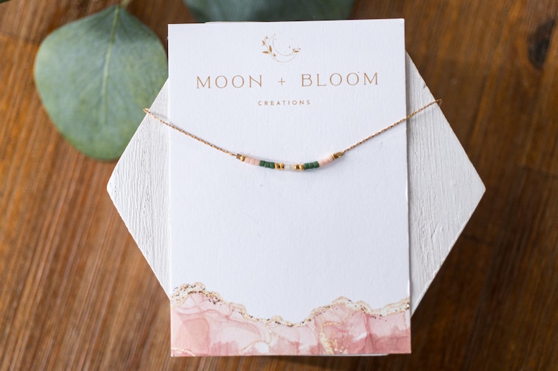 Puede incluir: Un delicado collar de cadena dorada con peque&ntilde;as cuentas rosas y verdes, presentado en una tarjeta blanca con el texto "MOON + BLOOM CREATIONS".