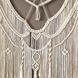 May include: Cream-colored macrame wall hanging with intricate knotted patterns and fringe. The design features braided sections, geometric shapes, and cascading vertical strands, creating a textured, bohemian aesthetic. The wall decor is mounted on a wooden dowel.