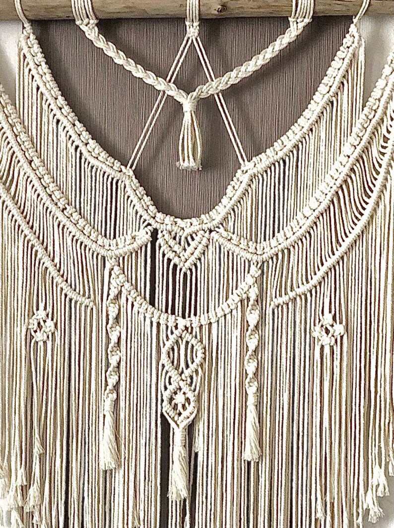May include: Cream-colored macrame wall hanging with intricate knotted patterns and fringe. The design features braided sections, geometric shapes, and cascading vertical strands, creating a textured, bohemian aesthetic. The wall decor is mounted on a wooden dowel.