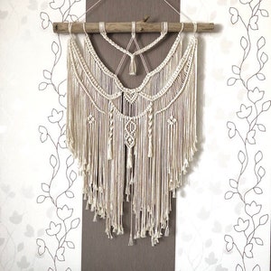 May include: Cream-colored macrame wall hanging. The handcrafted piece features intricate knotting patterns, a wooden dowel, and long, flowing fringe. The design includes geometric shapes and decorative tassels, creating a bohemian aesthetic. The background is a floral wallpaper.