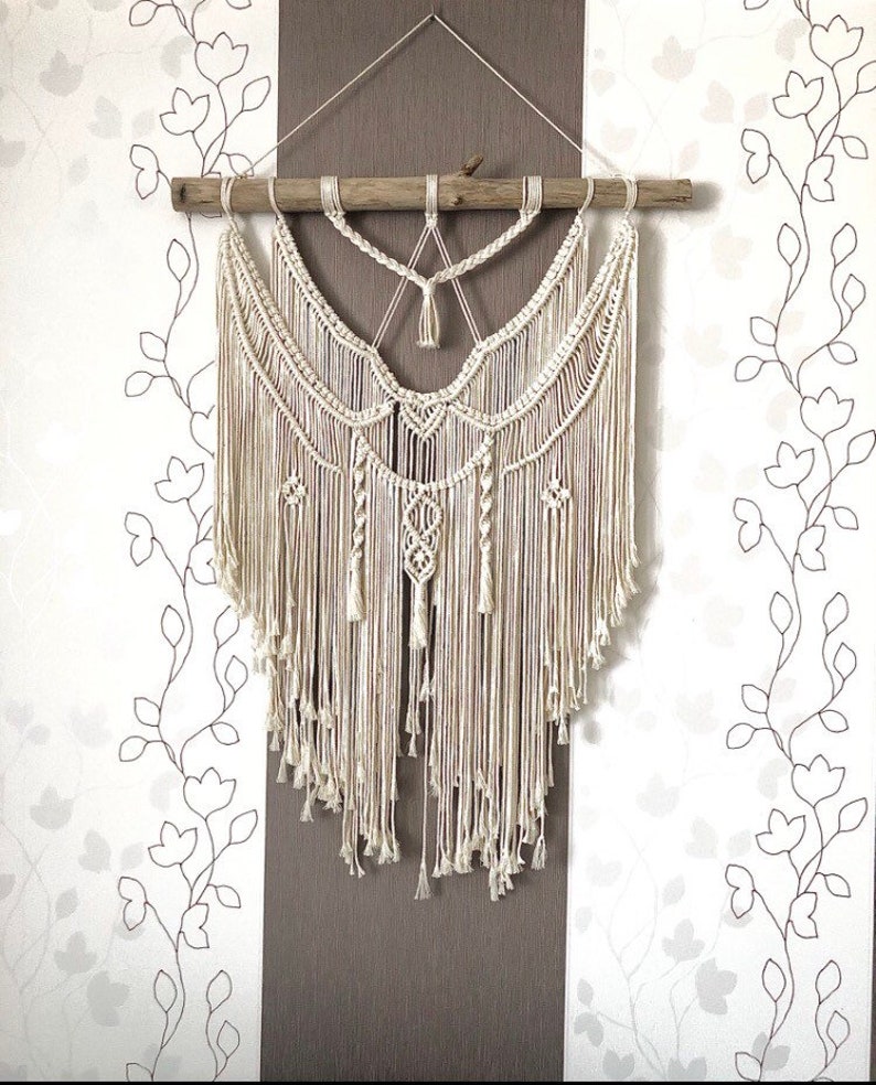 May include: Cream-colored macrame wall hanging. The handcrafted piece features intricate knotting patterns, a wooden dowel, and long, flowing fringe. The design includes geometric shapes and decorative tassels, creating a bohemian aesthetic. The background is a floral wallpaper.