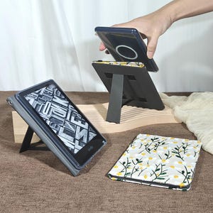 Może przedstawiać: Szara obudowa czytnika e-booków ze stojakiem, wyświetlająca czarno-biały obraz. Ręka trzyma podobną obudowę z magnetycznym okręgiem ładowania. W pobliżu leży etui z kwiatowym wzorem. Obudowy są przeznaczone do przechowywania czytników e-booków.