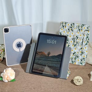 May include: A tablet with a black screen displaying the time 15:01, resting on a gray stand. A second tablet with a clear case and an Apple logo is visible. A third item is a white and yellow floral patterned case.
