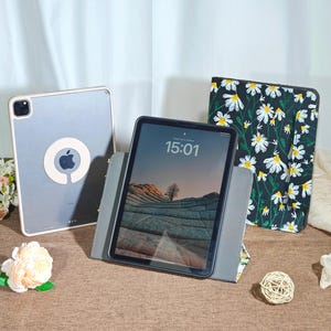Puede incluir: Se muestran tres tabletas con fundas. Una tableta está en una funda transparente con un logotipo de Apple. Otra tableta está apoyada en un soporte gris, mostrando una imagen de paisaje y la hora 15:01. Una tercera funda de tableta presenta un fondo negro con flores de margarita blancas.