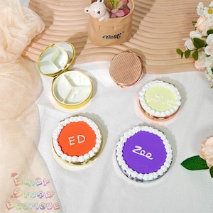 Peut inclure: Divers miroirs compacts ronds, avec des bords dorés et cuivrés. Certains miroirs ont un design en forme de gâteau avec du glaçage blanc et du texte. Un miroir est ouvert, révélant un intérieur divisé.