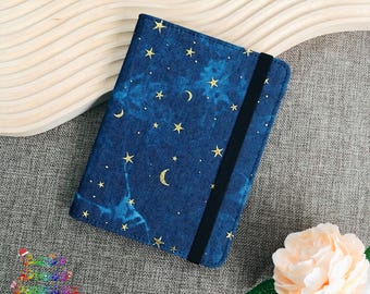 Könnte beinhalten: Ein blauer Notizbuchumschlag mit einem Muster aus goldenen Sternen und Mondsicheln. Der Umschlag hat einen schwarzen Gummibandverschluss.
