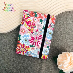Può includere: Una copertina per quaderno con motivo floreale e chiusura elastica nera. La copertina presenta una varietà di fiori rosa, rossi, blu e gialli su uno sfondo bianco.