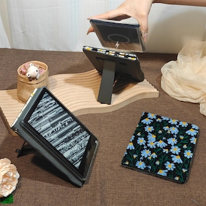 May include: A black e-reader case featuring a pattern of blue daisies with green stems on a black background. The case includes a built-in stand, a magnetic charging component, and a protective cover that folds to support the device in an upright position.